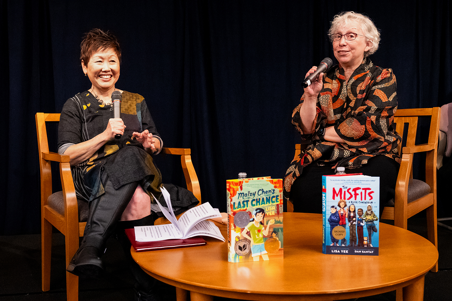 Two women sit side by side around a small circular table and both hold microphones. On the table in front of them sit two books by Lisa Yee: Maizy Chen's Last Chance and The Misfits