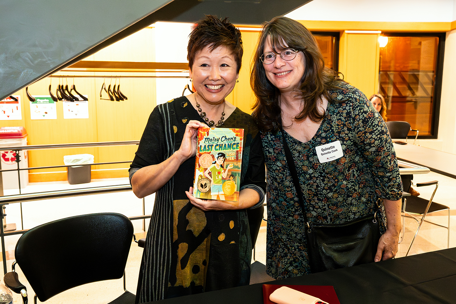 Two women sit side by side around a small circular table and both hold microphones. On the table in front of them sit two books by Lisa Yee: Maizy Chen's Last Chance and The Misfits