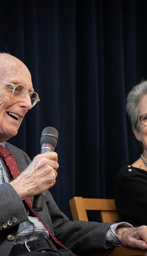 Man holds a microphone and talks while a woman next to him listens and smiles widely