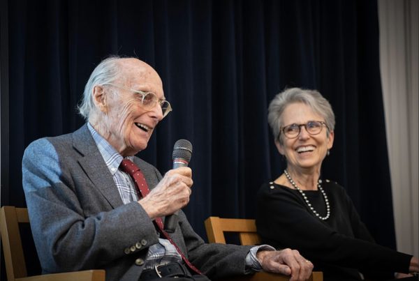 Man holds a microphone and talks while a woman next to him listens and smiles widely